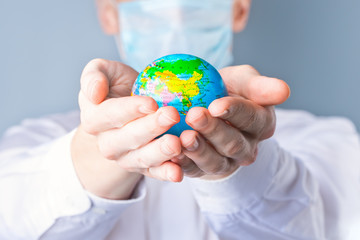 Close-up of a man's hands holding a small globe, a man in a medical mask out of focus, dressed in a white shirt, uniform on a gray background.  2020, the global economic crisis. Coronavirus, Covid-19
