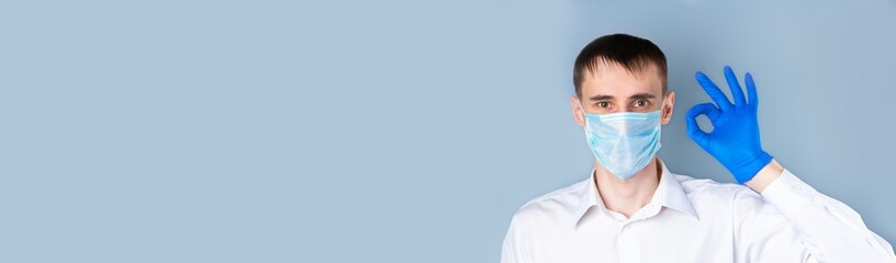 A man in a medical mask shows the OK gesture in a blue medical glove, wearing a white shirt on a gray background, left empty space for text.  Banner. Coronavirus, Covid-19