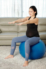 Smiling young pregnant woman sitting on fitness ball and outstretching arms when doing balance exercise