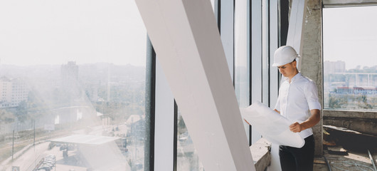 Caucasian businessman holding a building plan and analyzing it while posing near a big window with a helmet