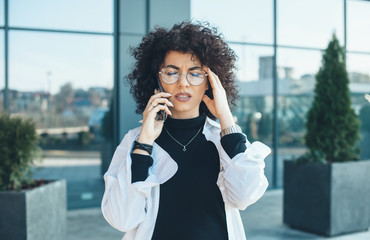 Thoughtful caucasian lady with curly hair and glasses touching her hair and posing outside during a business phone talk