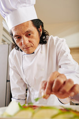 Mature Vietnamese man in chef coat and hat garnishing dish with greens