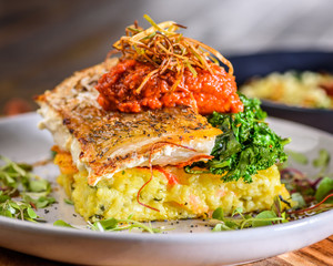 A serving of Fish of The Day, Snapper with Crispy Paella Cake, Wilted Kale, Spinach, Dukkah Tomato Config Topped with Crispy Leek Straws
