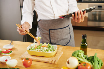 Cropped image of cook checking recipe on tablet computer and mixing salad with chopsticks