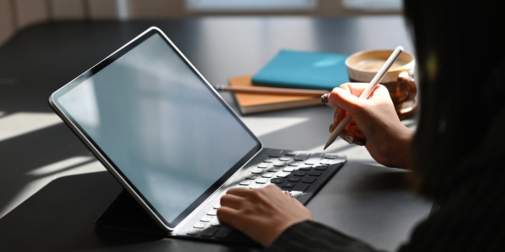 Cropped image of businesswoman holding a stylus pen while using a computer tablet and sitting at the black working desk over comfortable office as background.