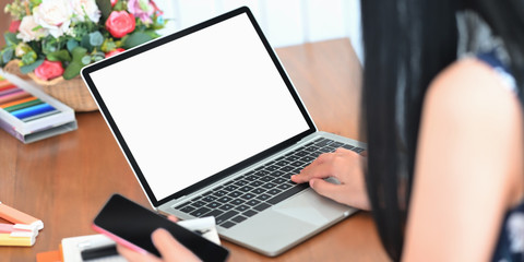 Cropped image of beautiful woman holding a smartphone in hand while using a computer laptop and sitting at the wooden working desk over comfortable living room as background.