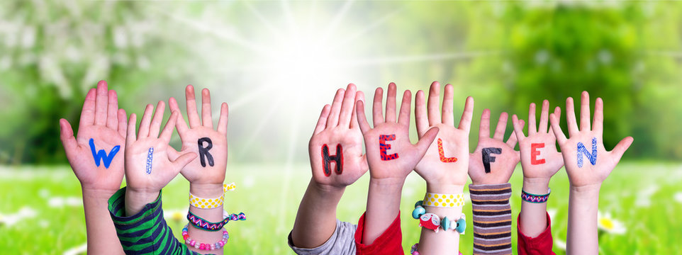 Kids Hands Holding Colorful German Word Wir Helfen Means We Help. Sunny Green Grass Meadow As Background