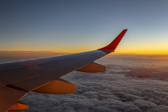 Aircraft wing with fluffy clouds below and orange horizon during sunset