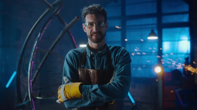 Young Professional Fabricator in Safety Glasses and Apron Gently Smiles at the Camera and Crosses Arms. Authentic Artist Wearing Work Clothes in a Metal Workshop. Sparks Flying on the Background.