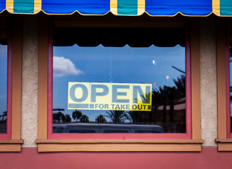 Yellow sign with black letters in the window at restaurant stating they are Open For Take Out . Awning in visible as well as palm trees reflected in window