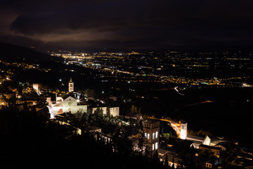 Aerial view of Assisi town (Umbria, Italy) and valley at night, with city lights and Santa Chiara church