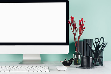 Fresh home workspace with blank computer monitor, black stationery, books, red flowers, keyboard, mouse, coffee cup in green mint menthe interior on white wood desk, closeup.