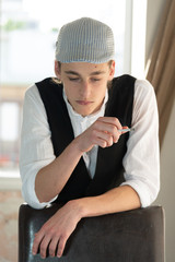 A young man in a cap and vest smokes by the window