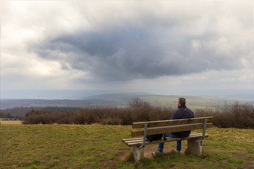 A hiker with a backpack sits on a wooden bench. The place is called Köterberg. It is the highest mountain in eastern Westphalia in Germany. It is a winter day with impressive clouds.