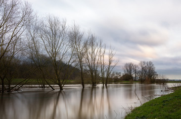 An atmospheric German river landscape in winter in the east of North Rhine-Westphalia near the city of Höxter. A long exposure with soft water and moving clouds. Trees are submerged in the flood.