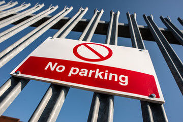 Low angle view, of a red & white No Parking sign attached to a metal security fence, at an industrial trading estate in Colliers Wood, London, UK