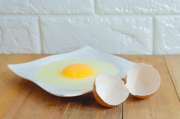 Eggs hammered on a white plate with an egg shell on the side, placed on a wooden table in the kitchen with a brick background.
