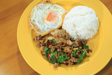 Rice topped with stir-fried pork and basil, fried egg on a yellow plate on a wooden table