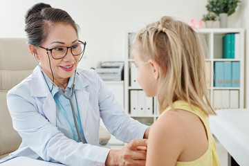 Obraz premium Smiling mature Asian female pediatrician checking heartbeat of little patient