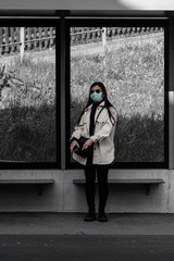 young woman standing at a bus stop wearing a breathing mask