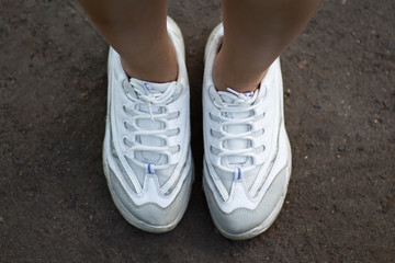 Two slim feet in white sneakers standing on the ground. Top view