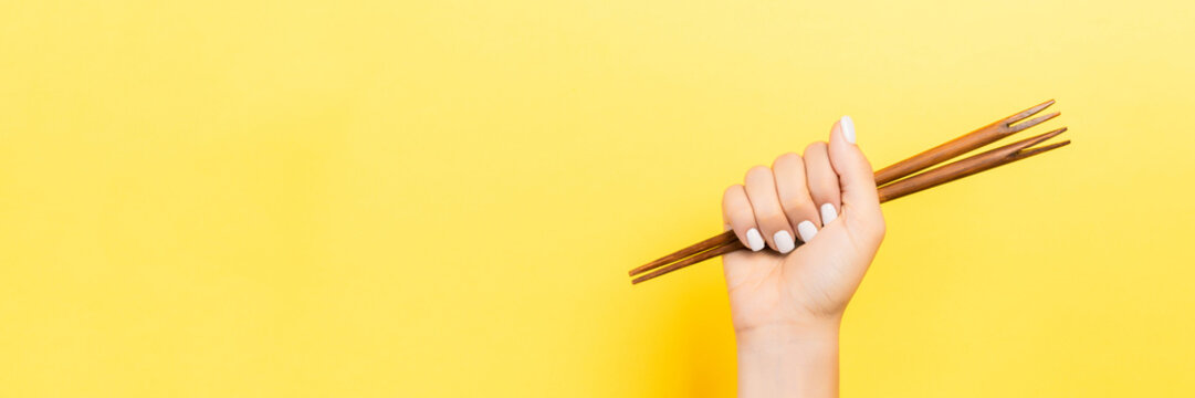 Cropped image of female hand holding chopsticks in fist on yellow background. Asian food concept with copy space