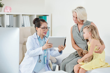 Obraz premium Mature Asian pediatrician showing results of medical tests on screen of tablet computer to mother of little girl