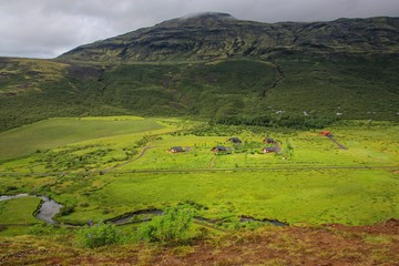Fototapeta premium small houses are located in a valley at the foot of green hills, near the winding river flows, the sky is overcast, the nature of Iceland, a summer day
