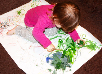 a small child draws with a gouache brush on a white sheet of paper on the floor. The view from the...