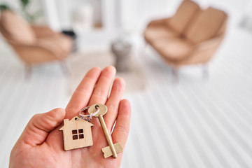Men hand holding key with house shaped keychain. Modern light lobby interior. Mortgage concept. Real estate, moving home or renting property.