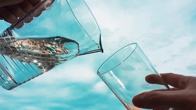 Llenando un vaso de agua con una jarra a c&aacute;mara lenta con un cielo de nubes al fondo.