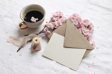Spring still life scene. Greeting card mockup, envelope, cup of coffee on white linen table cloth. Vintage feminine styled photo. Floral composition with pink sakura, cherry tree blossoms, top view.