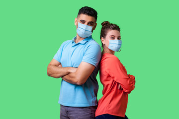 Reliable friendship. Cheerful young couple of friends with surgical medical mask standing back to back with crossed hands and looking at camera . isolated on green background, indoor studio shot