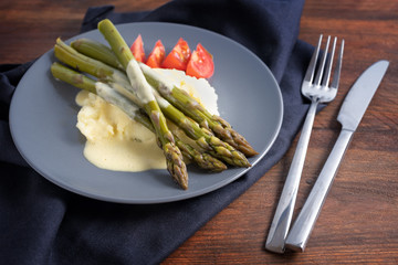 Green asparagus on mashed potatoes with light hollandaise sauce and tomatoes on a blue plate, napkin and cutlery on a dark rustic wooden table