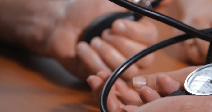 Close-up detail shot of hands of an elderly couple. Husband takes care of his wife by measuring her blood pressure using a mechanical tonometer on a brown wooden table at home. 4k 50fps slow motion