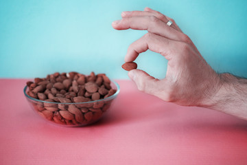 Close up man hand holds one almond nut on a colored background - blue and pink table next to a glass plate