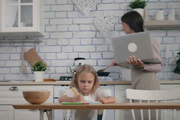 Mother and her five year old daughter on the kitchen while self isolation in case of quarantine. Woman wearing apron work with laptop during cooking, her daughter drawing at the table. Stay home remot