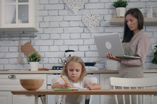 Mother and her five year old daughter on the kitchen while self isolation in case of quarantine. Woman wearing apron work with laptop during cooking, her daughter drawing at the table. Stay home remot