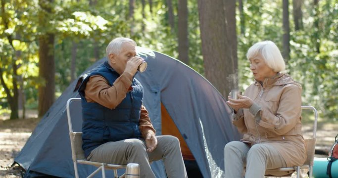 Old Caucasian happy couple of hikers resting in forest and drinking tea. Male and female tourists sipping hot drink at tent. Senior man and woman having rest in wood. Camping and tourism concept.