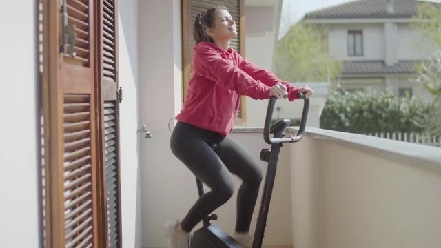 Cheerful girl pedaling on exercise bike on balcony
