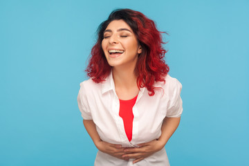 Laughter, positive emotions. Portrait of happy woman with fancy red hair in white shirt holding her belly while laughing out loud after hearing joke. indoor studio shot isolated on blue background