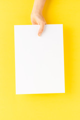 Overhead shot of woman’s hand holding blank paper sheet on yellow table. Close up