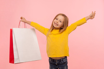 Fototapeta premium Portrait of joyful carefree little girl holding large packages and smiling happily, excited about shopping in children store, black Friday discounts. indoor studio shot isolated on pink background