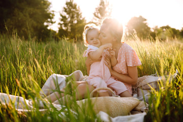 Mother and little daughter playing together in a summer park at sunset. Happy mom with small child  having fun on sunny field. Kisses and hugs. The concept of a happy family. 