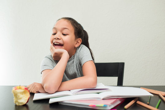 Positive little girl doing homework at home looking a sweet apple. School girl rests her head on her hand on a white background.