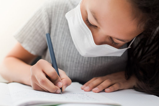 Schoolgirl doing writing on the notebook wearing a medical mask to protect from coronavirus infection during the quarantine. Concept of studying at home.