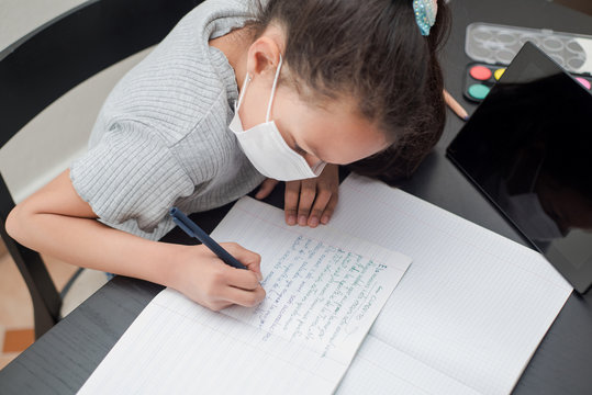 Pretty little girl taking notes on the notebook following online lessons on the tablet. Schoolgirl wears a protective mask during the coronavirus covid19 infection.