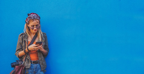 Young girl standing by the blue background holding a smartphone - A hipster stylish girly smiling and checking out social media