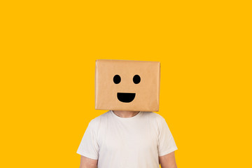 Hombre con caja de cartón en la cabeza con una sonrisa en la cara sobre fondo amarillo liso brillante. Vista de frente. Copy space - obrazy, fototapety, plakaty