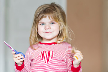 Little cute toddler girl making experiment with scissors and cutting hairs. Funny baby child cuts her pony herself at home. Trouble